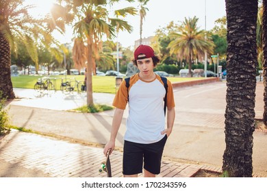 Male in sportswear standing with skateboard on empty road with palm trees in sunlight while looking at camera and holding one hand in pocket - Powered by Shutterstock