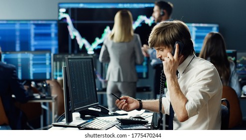 Male stock trader working at stock exchange office and talking phone on background of his business team and multiple monitors showing data, ticker numbers and graphs.  - Shutterstock ID 1775470244