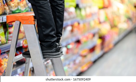 Male Storekeeper Legs Standing On Aluminum Stair Putting Products On Shelves In Grocery Store Or Supermarket. Worker Filling Foods Stock For Groceries Business.