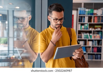 A male student, clad in a yellow tee, thoughtfully uses his digital tablet while leaning against the glass pane of a library window, a leather bag slung over his shoulder. - Powered by Shutterstock