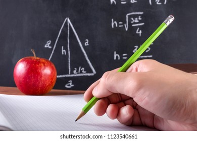 Male student hand writes in notebook with green wooden pen. Red apple and blackboard is blurred in background. Classroom and education concept. Close up, selective focus - Powered by Shutterstock