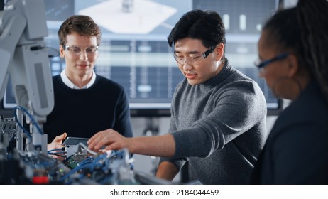 Male Student Holding A Tablet And Pointing At Robotic Hand While Working On Project With Asian Classmate And Black Teacher. Modern Technologies, Science And Developing At University Concept.