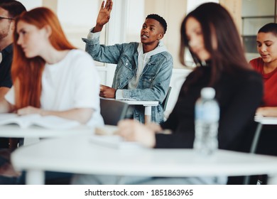 Male Student Sitting In The Class And Raising Hand To Ask A Question During Lecture. College Student Asking A Query To The Lecturer In Classroom.