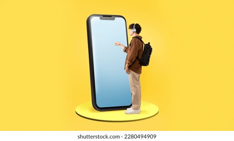 Male Student Using Large Smartphone With Blank Screen Standing With Backpack On Yellow Studio Background. Guy Touching Phone Touchscreen Advertising Educational App. Panorama, Full Length - Shutterstock ID 2280494909