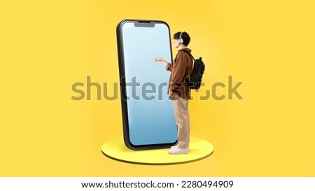Male Student Using Large Smartphone With Blank Screen Standing With Backpack On Yellow Studio Background. Guy Touching Phone Touchscreen Advertising Educational App. Panorama, Full Length