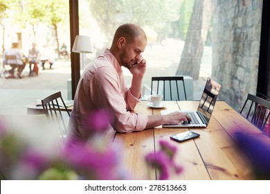 Male Student Work On Computer Touch Pad While Talking On Smart Phone, Young Business Man Use Laptop Sitting At Wooden Table Of Modern Coffee Shop, Freelancer Working On Notebook At Hipster Loft Space