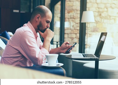 Male Student Work On His Computer While Chatting On Smart Phone, Young Business Man Use Laptop Sitting At Sofa Of Modern Coffee Shop Or Hotel Hall, Freelancer Working On Notebook At Hipster Loft Space