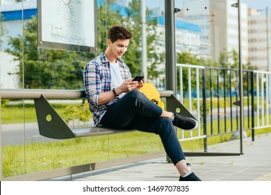 Male student wth smart phone wearing plaid shirt sitting on bench while waiting for transport at tram or bus station. City public transport infrastructure, citizen and tourists transportation concept