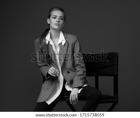 Male style dressed fashion female model. Fashionable woman in blouse, blazer, trousers and boots sitting at high cjair and posing against gray background. Stylish fashionable blonde girl