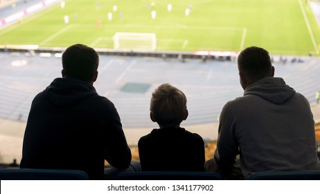 Male Supporters And Child Watching Football Match At Stadium, Discussing Game