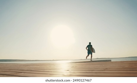 A male surfer runs along a pier on a lake against the sun, holding a water board. A male surfer walks along the pier with a small surfboard in his hands. Summer water sports - Powered by Shutterstock