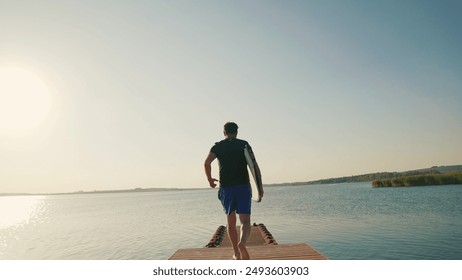 A male surfer runs along a pier on a lake against the sun, holding a water board. A male surfer walks along the pier with a small surfboard in his hands. Summer water sports - Powered by Shutterstock