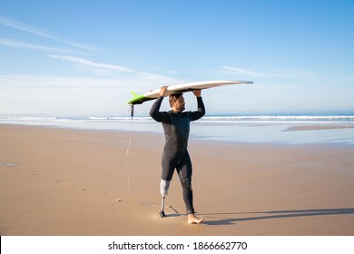 Male surfer wearing artificial limb and wetsuit, walking on beach, carrying surfboard overhead. Full length. Artificial limb and active lifestyle concept - Powered by Shutterstock