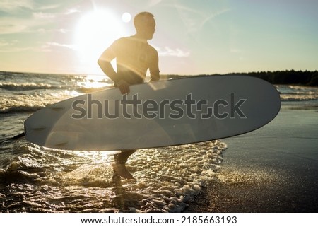 A male surfer in a wetsuit riding in the sea on a sunny day at sunset, holding surfing in his hands