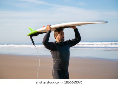 Male Surfer In Wetsuit Walking On Beach And Carrying Surfboard Overhead. Medium Shot. Surfing And Active Lifestyle Concept