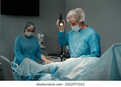 Male surgeon adjusting lamp before surgical operation while standing by operating table with patient covered with napkin - Powered by Shutterstock