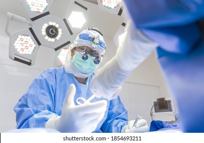 Male Surgeon In Blue Surgical Gown Suit Inside Modern Cardio Thoracic Operating Room With Loupe Magnificent Glasses. Doctor Perform Heart Surgery. Selective Focus At Surgeon 's Eyes. Medical Concept.