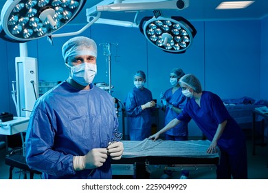 Male surgical doctor in blue scrub suit with surgical scissors in hands standing inside operating room with surgery team - Powered by Shutterstock