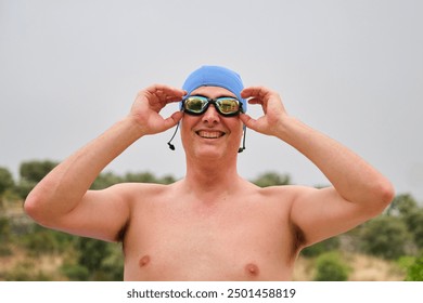 Male swimmer with blue swim cap smiling while adjusting goggles outdoors before a swimming competition - Powered by Shutterstock