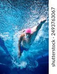 Male swimmer, seen underwater, propels himself forward with powerful strokes, wearing blue swim trunks, goggles and cap. Concept of professional sport, competition, motion. Underwater shot.