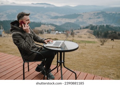 Male talking on phone outdoors. Man work on a laptop sitting table with great view mountains. Concept remote work or freelance lifestyle. Internet 5G. Workplace in country on backyard house in morning - Powered by Shutterstock