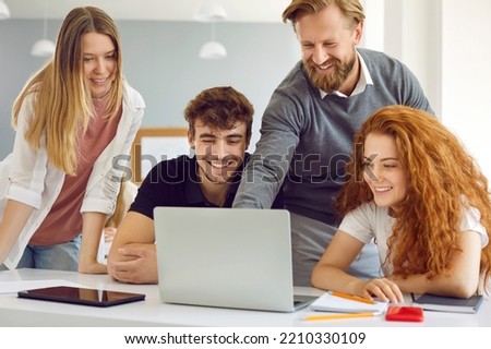 Male teacher helps group of college students working on laptop during computer lesson. Positive teacher points to laptop screen and explains something to three smiling young people sitting at desk.