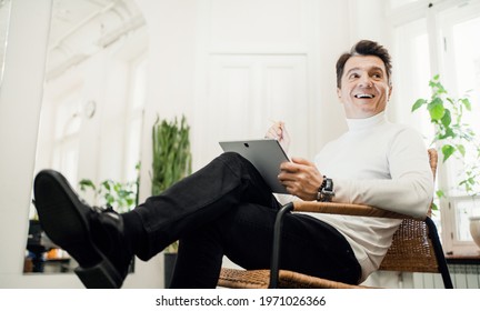 A Male Teacher At A Music School. Sitting On A Chair And Holding A Tablet With A Lesson Plan. An Adult Creative Person.