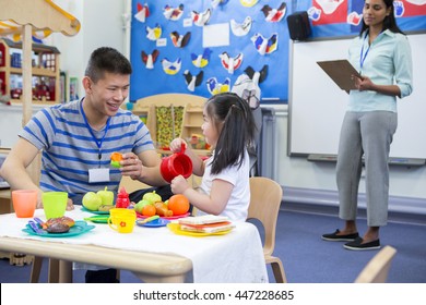 Male teacher playing in a toy kitchen with a nursery student. There is a teacher in the background with a clipboard who is watching them.  - Powered by Shutterstock