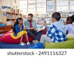 Male teacher reading a book to students while sitting in school library. Librarian read a fairy tale to a group of multiethnic primary schoolchildren. Mid adult man at elementary school reading story.