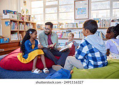 Male teacher reading a book to students while sitting in school library. Librarian read a fairy tale to a group of multiethnic primary schoolchildren. Mid adult man at elementary school reading story.