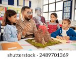 Male teacher teaching to a group of school children about volcanos at science project in school. Group of students understanding volcanic eruption in class with teacher while observing a volcano model