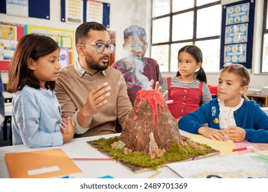 Male teacher teaching to a group of school children about volcanos at science project in school. Group of students understanding volcanic eruption in class with teacher while observing a volcano model