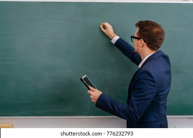 Male Teacher Writing On A Blank Green Chalkboard With Copy Space While Holding A Tablet Computer In His Other Hand