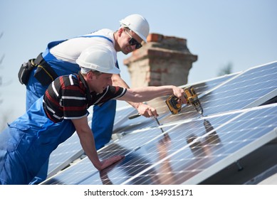 Male team workers installing stand-alone solar photovoltaic panel system using screwdriver. Electricians mounting blue solar module on roof of modern house. Alternative energy sustainable concept