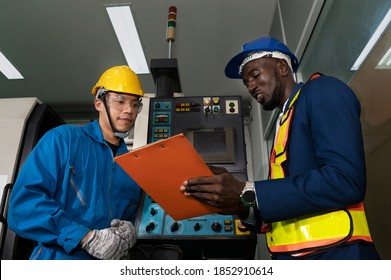 Male Technician Inspector Discusses With Male Factory Mechanic Expert In Front Of A Machine Control Panel In Factory Maintenance Office With Protective Glasses, Gloves And Helmet In Factory Uniform