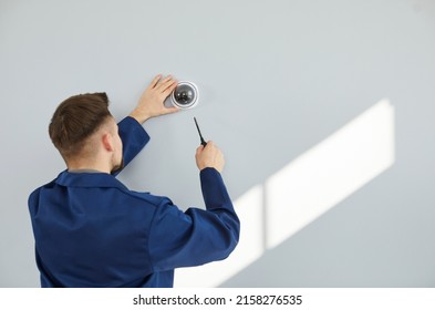 Male Technician Installing Surveillance Camera On Light Copy Space Wall. Back View Of Repair Service Worker Using Screwdriver To Fit Screws And Adjust Wall Mounted CCTV Security Dome Cam Inside House