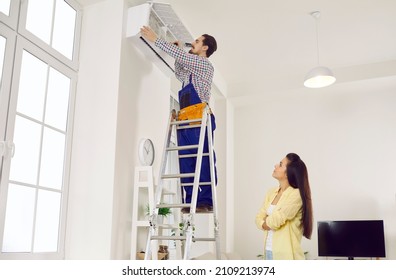 Male Technician Repair Air Conditioner In Female Customer Home. Man Engineer Or Repairman On Ladder Fixing Conditioning Device At Client Apartment. Electrician Work With Gadget. Engineering Company.