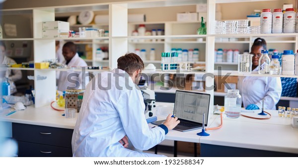Male technician sitting\
at a table in a lab looking through a microscope and entering data\
into a laptop 