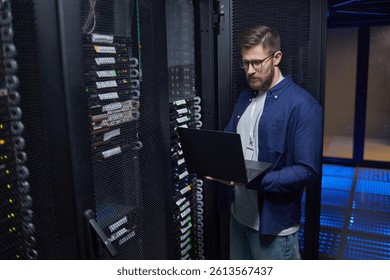Male IT technician standing in modern server room holding laptop, checking network equipment and ensuring proper functionality. High-tech environment featuring server racks and data cables