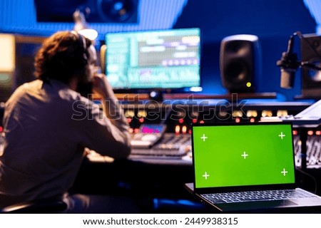 Male technician working on editing tracks next to greenscreen on laptop, operates buttons and knobs in control room. Audio engineer using faders and other technical equipment, mix and master.