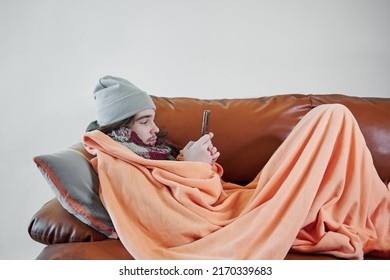Male teenager freeze at home and try to keep warm covered under warm plaid on sofa with smartphone. Power crisis concept, problem with heating - Shutterstock ID 2170339683
