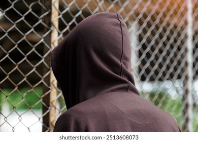  male teenager wearing a brown hoodie stood facing a steel mesh fence. Both hands cling to the fence, showing the back.                               - Powered by Shutterstock