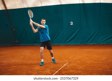 A male tennis player in a blue shirt and black shorts is preparing to hit a backhand shot on an indoor clay court. The court is marked with lines and has a green backdrop. - Powered by Shutterstock