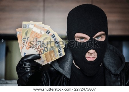 Male thief with balaclava on his head holding a handful of euro bills in front of his eyes. the concept of crime, theft and illegal business, money laundering