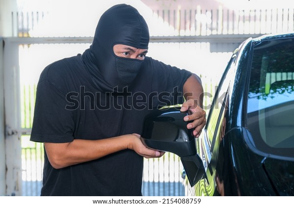 A male thief wearing a balaclava is trying to\
break a car\'s rearview\
mirror.