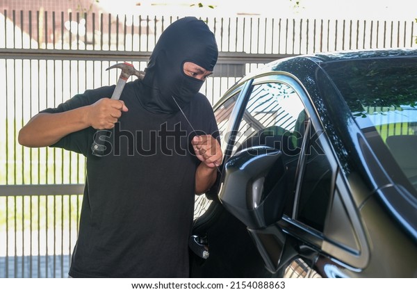 A male thief wearing a balaclava is trying to\
break a car window with a\
hammer.