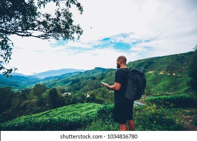 Male tourist with black backpack holding smartphone with online navigator and enjoying high green hills and landscapes during expedition in faraway asia places.Hiker during wanderlust in mountaints