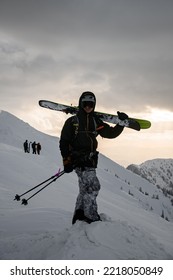 Male Tourist In Black Ski Suit And With Backpack With Skies On His Shoulders On Snow-covered Mountain. Ski Touring And Freeride Concept