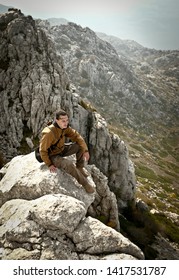 Male Tourist (brown Membrane Jacket, Grey Cargo Pants) Sitting On Top Of Mountain. Several Mountain Ridges Behind Him With Rocks And Green & Yellow Moss. Vertical Perspective. Croatia Natural Reserve