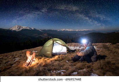 Male tourist have a rest in his camp at night. Man with lighting headlamp sitting near campfire and tent, looking to the camera under beautiful sky full of stars and milky way